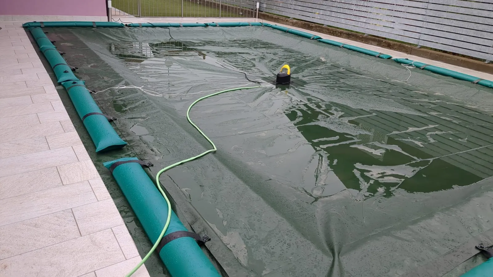 Piscina chiusa per l’inverno: telo verde fissato dai salsicciotti lungo il bordo e una pompa che toglie l’acqua piovana