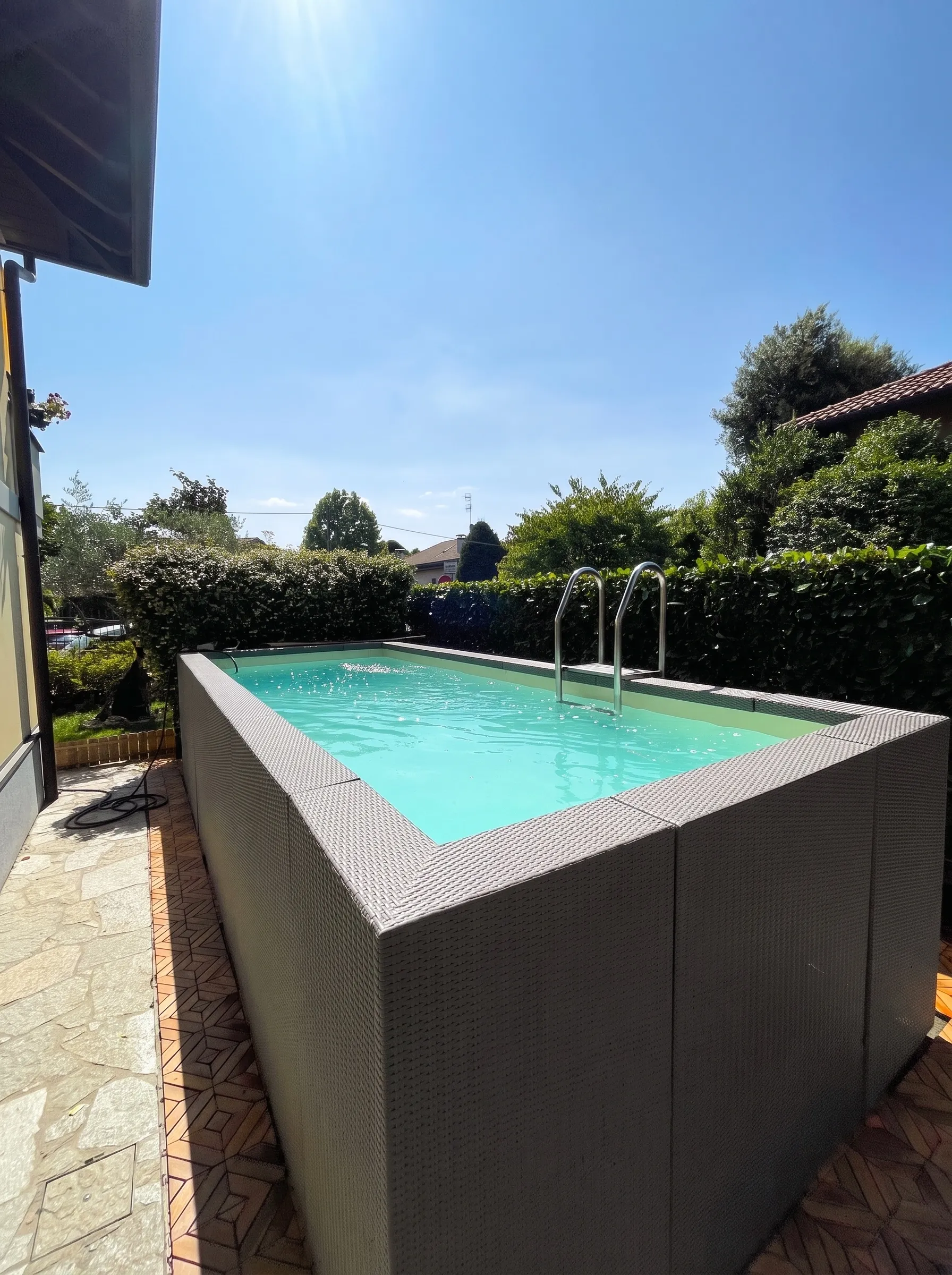 Piscina fuori terra con rivestimento effetto rattan su una pavimentazione, siepe e scaletta in acciaio