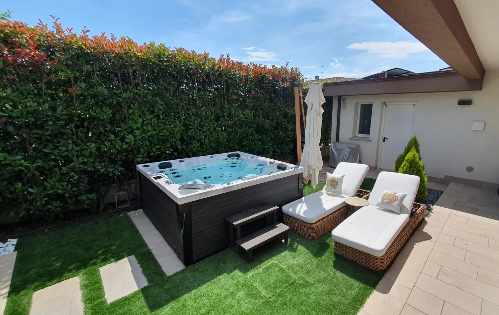 Spa idromassaggio installata in giardino accanto a due lettini, con siepe e ombrellone chiuso